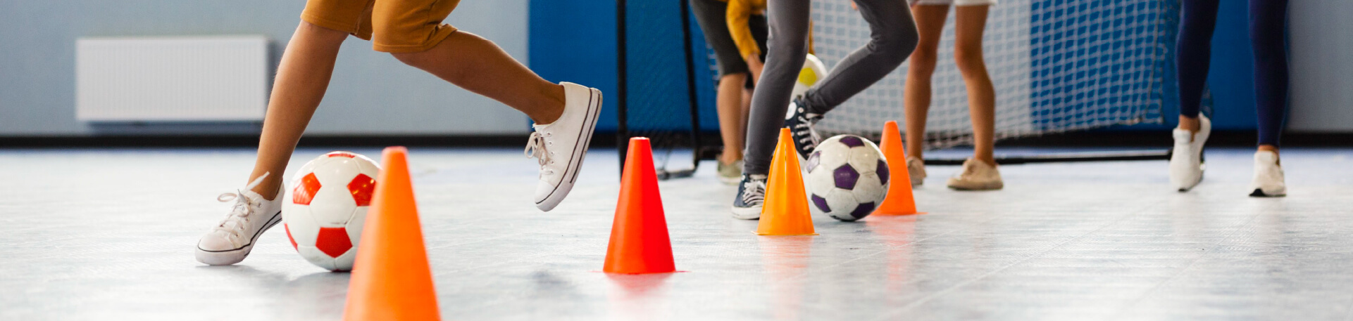 Crianças a realizar exercícios de condução de bola com obstáculos em interior, usando bolas de futebol e cones coloridos, num espaço desportivo com baliza ao fundo.