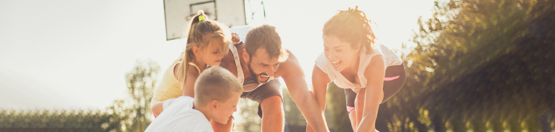 Família a brincar com uma bola de basquetebol num campo exterior, com expressões de alegria e envolvimento de todos os elementos.