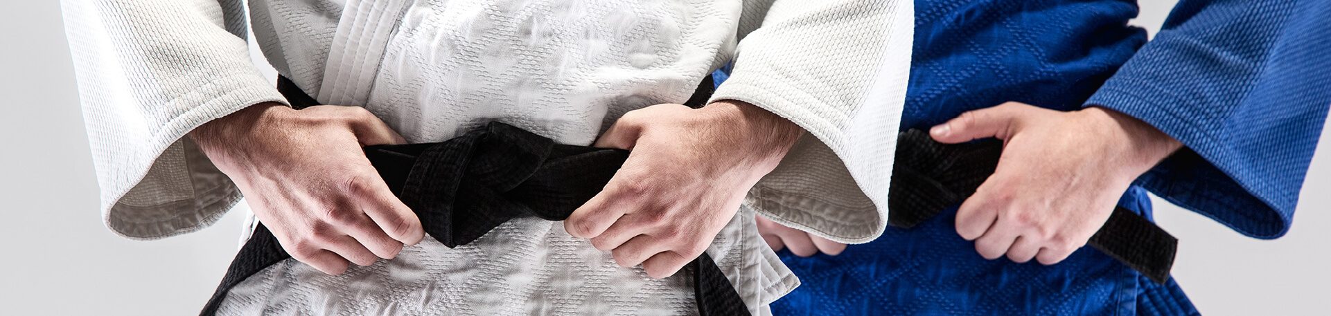 Dois praticantes de artes marciais com quimonos, um branco e outro azul, com cintos pretos, focando-se na zona do tronco e das mãos a ajustar o cinto.