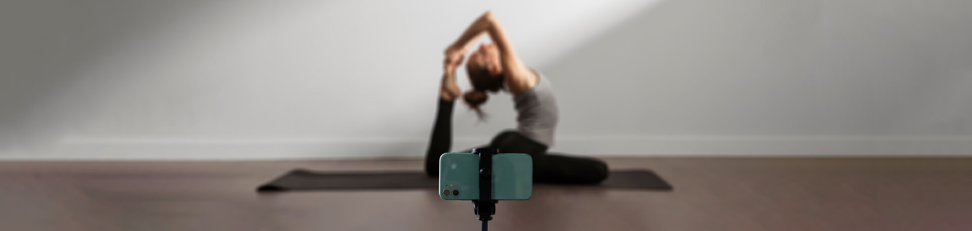 Smartphone montado num tripé em primeiro plano, com uma mulher ao fundo a praticar yoga numa sala, numa posição de alongamento com uma perna esticada e as mãos a segurar o pé por trás da cabeça.
