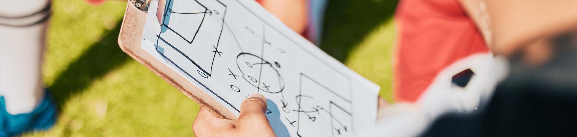 Pessoa segura uma prancheta com um esquema tático desenhado, rodeada por jogadores com equipamento de futebol vermelho e branco, sentados ou ajoelhados sobre relvado.