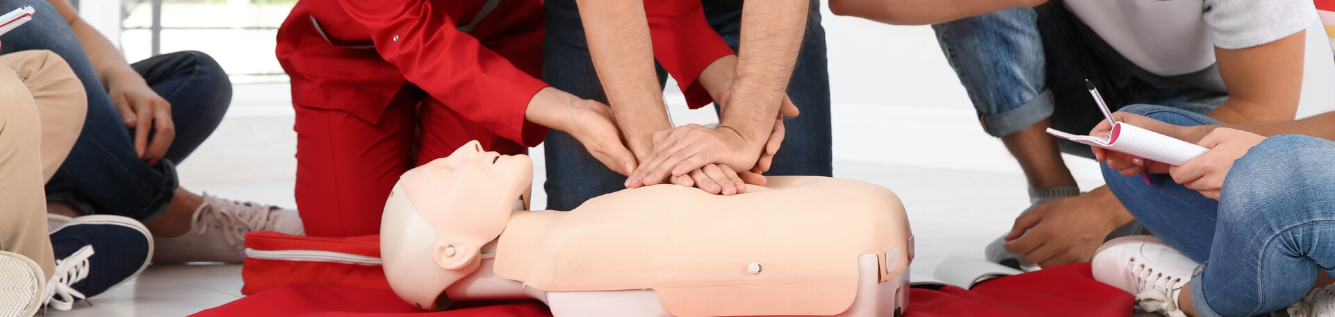 Homem a realizar compressões torácicas num manequim de reanimação, sob orientação de uma formadora, enquanto outros participantes observam atentamente durante uma sessão de primeiros socorros.