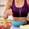 Mulher com roupa de desporto roxa e auriculares, numa cozinha, a preparar uma taça azul com fruta, colocando um pedaço de morango por cima. Na mesa estão também outras frutas cortadas e inteiras.