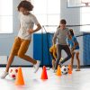 Crianças a realizar exercícios de condução de bola com obstáculos em interior, usando bolas de futebol e cones coloridos, num espaço desportivo com baliza ao fundo.