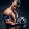Homem musculado sem camisola a segurar um haltere com o braço direito, usando uma braçadeira de oclusão no braço, num ambiente escuro com fundo neutro.