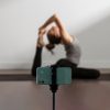 Smartphone montado num tripé em primeiro plano, com uma mulher ao fundo a praticar yoga numa sala, numa posição de alongamento com uma perna esticada e as mãos a segurar o pé por trás da cabeça.