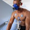 Homem a realizar um teste de esforço com máscara de medição de consumo de oxigénio e vários elétrodos colocados no peito, ligado a equipamento de monitorização fisiológica.