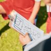 Pessoa segura uma prancheta com um esquema tático desenhado, rodeada por jogadores com equipamento de futebol vermelho e branco, sentados ou ajoelhados sobre relvado.