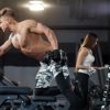 Homem sem camisa realiza exercício de remada unilateral com haltere, apoiado num banco de treino; ao fundo, mulher treina com halteres junto ao suporte.
