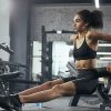 Mulher a realizar exercícios de tríceps num banco de musculação, vestida com roupa desportiva preta, num ginásio equipado com máquinas de musculação ao fundo.