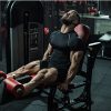 Homem musculado a treinar numa máquina de extensão de pernas no ginásio, com expressão de esforço e foco, vestindo equipamento desportivo preto.