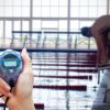 Cronómetro em primeiro plano junto a piscina olímpica, com nadador em posição de partida.