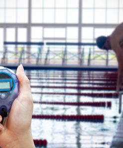 Cronómetro em primeiro plano junto a piscina olímpica, com nadador em posição de partida.